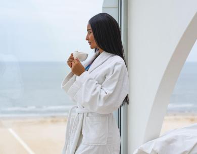 Woman in white robe drinks coffee with sea view.