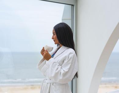 Donna in accappatoio bianco beve caffè con vista mare.