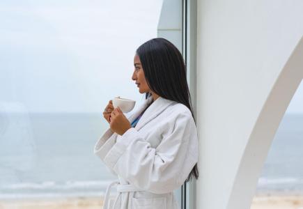 Femme en peignoir blanc boit du café avec vue sur la mer.