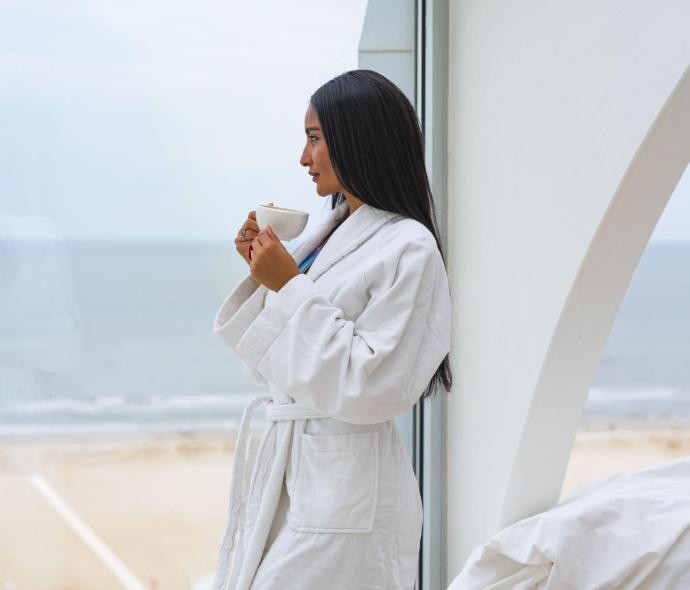 Woman in white robe drinks coffee with sea view.