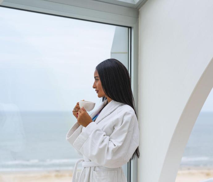 Donna in accappatoio bianco beve caffè con vista mare.